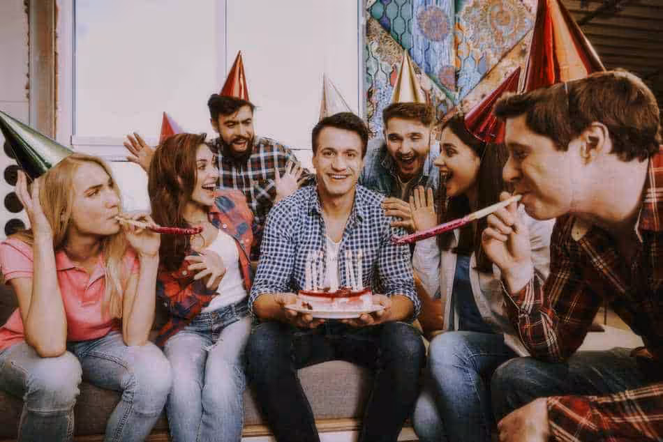 Eine Gruppe von sechs Personen mit Partyhüten feiert mit einem Kuchen. Eine Person hält den Kuchen mit brennenden Kerzen, während andere in Partytröten blasen und lächeln. Sie sitzen drinnen auf einem Sofa, während ein DJ im Hintergrund die festliche Stimmung für eine unvergessliche Feier anstimmt.